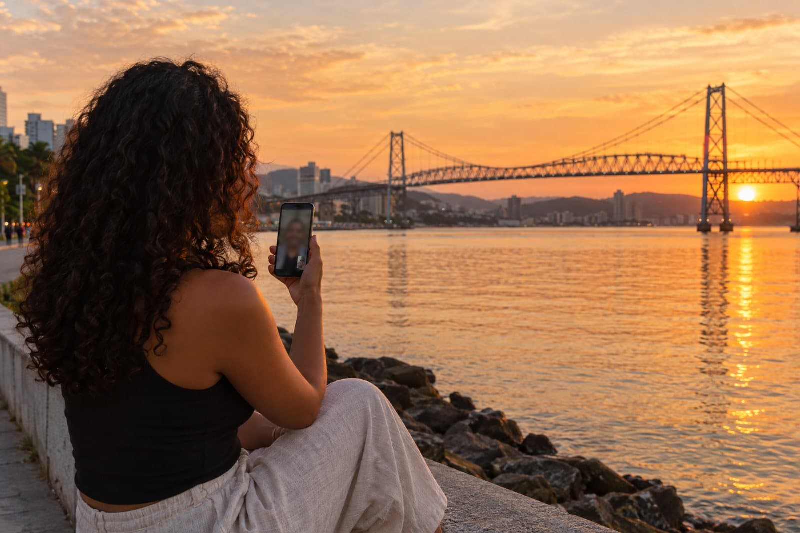Pessoa em videochamada pelo celular na Beira-Mar Norte, em Florianópolis, com a Ponte Hercílio Luz ao fundo, no pôr do sol