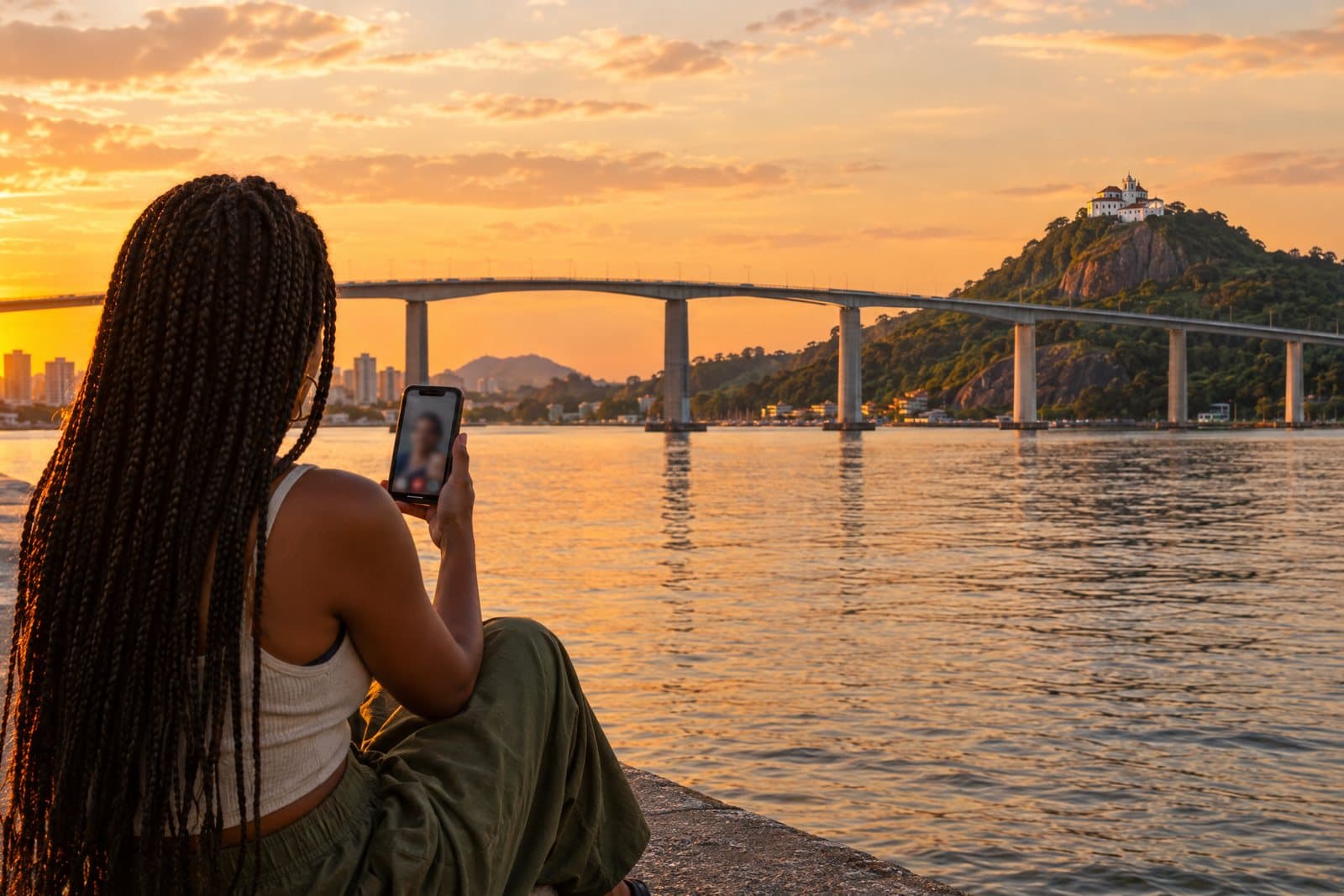 Pessoa em videochamada pelo celular na orla de Vitória, com a Terceira Ponte e o Convento da Penha ao fundo, no fim da tarde